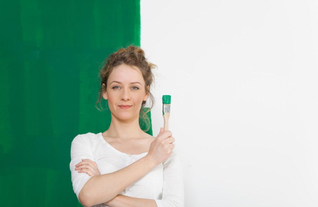 Frau mit grünem Pinsel steht selbstbewusst vor gestrichener Wand