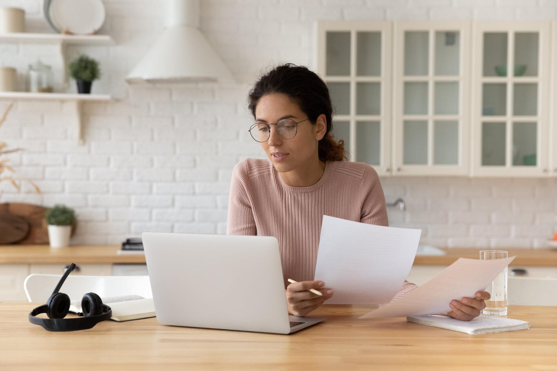 Frau sitzt am Schreibtisch und hält Finanzdokumente vor dem PC-Bildschirm, um Daten zu überprüfen und Angebote zu vergleichen.