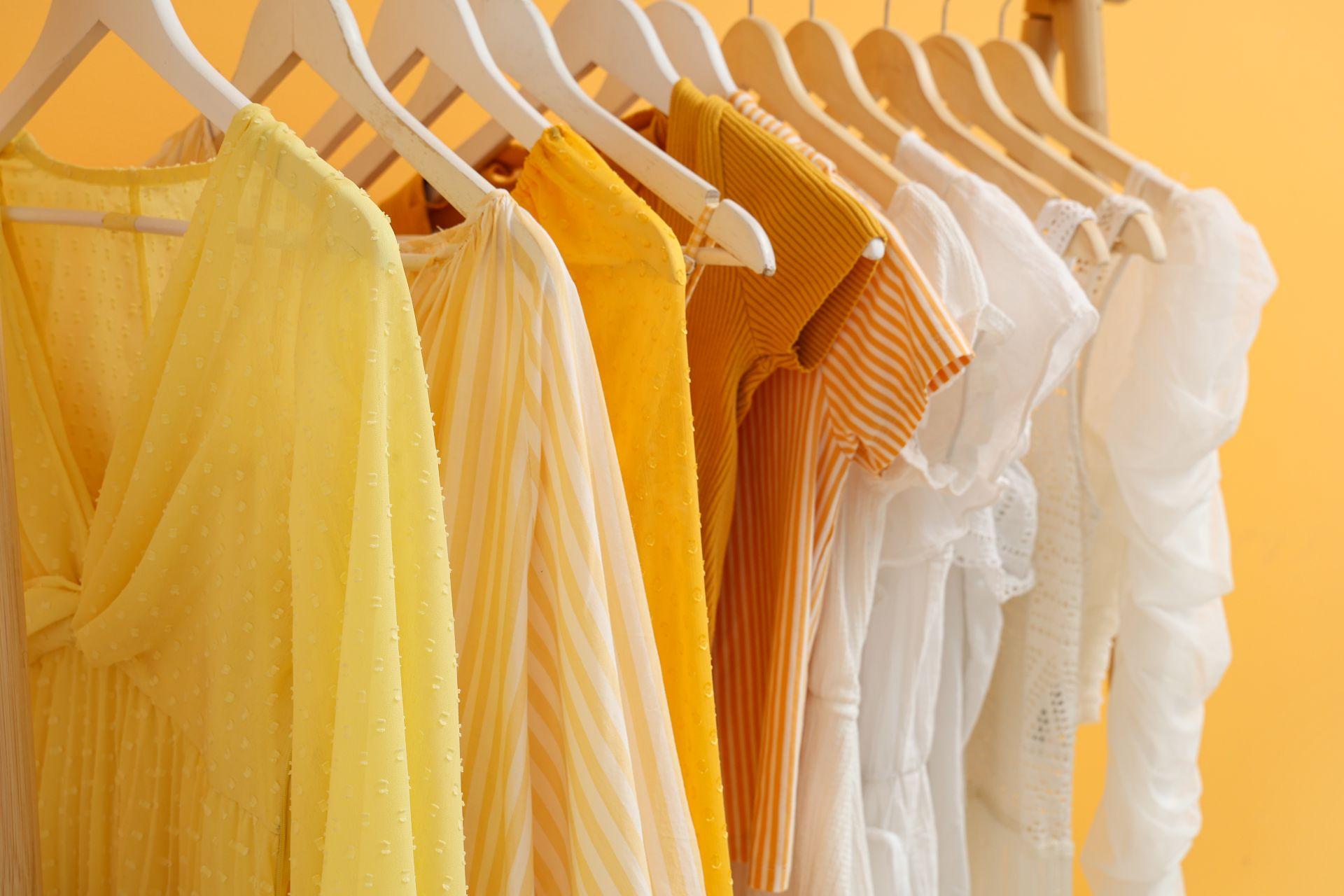 musselin kleidung und weitere leichte sommermaterialien in gelb und weiss ordentlich auf kleiderbuegeln vor warmem hintergrund